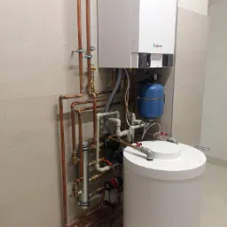 Nowoczesny, biały piec gazowy marki Viessmann z niebieskim zbiornikiem wyrównawczym i białym zasobnikiem ciepłej wody, połączony miedzianymi rurami w instalacji hydraulicznej w pomieszczeniu...