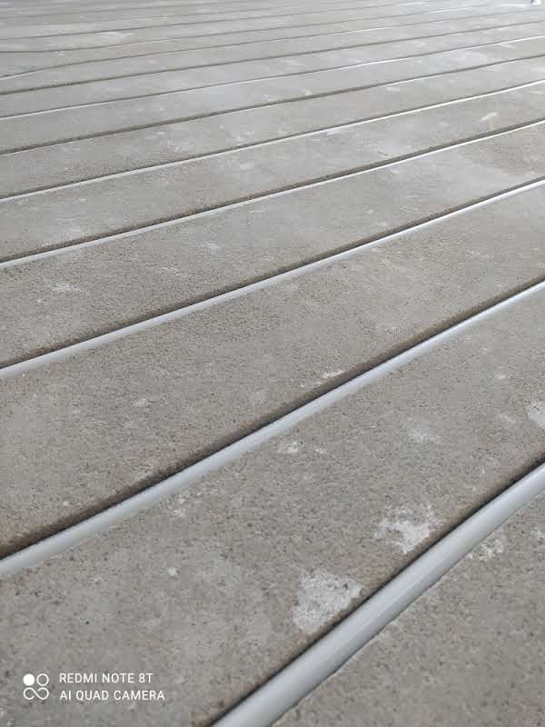 Szare panele podłogowe z widocznymi, jasnymi rurkami między nimi, ułożone równolegle, sfotografowane pod kątem.