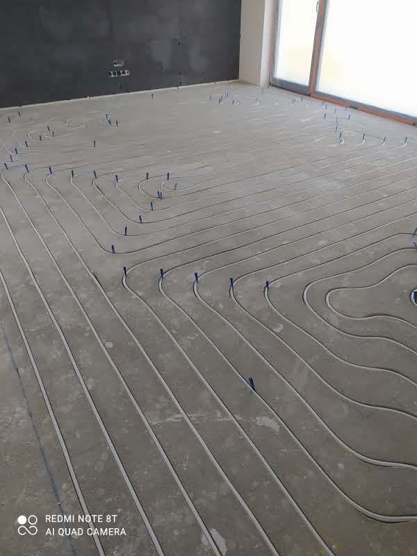 Szare rury ogrzewania podłogowego ułożone w pętle na surowym betonie, zabezpieczone niebieskimi uchwytami, widoczne okno i fragment ściany w tle.