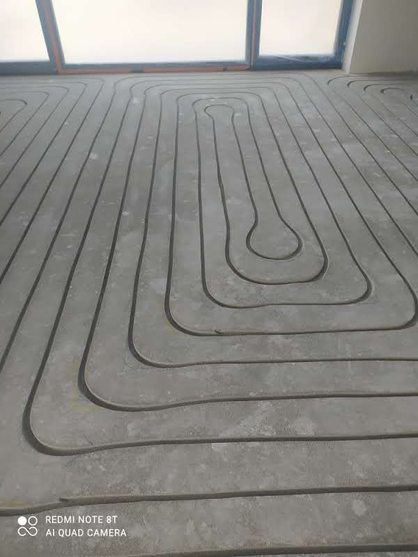 Ułożone rury ogrzewania podłogowego zatopione w szarej wylewce betonowej, widoczne okno w tle.