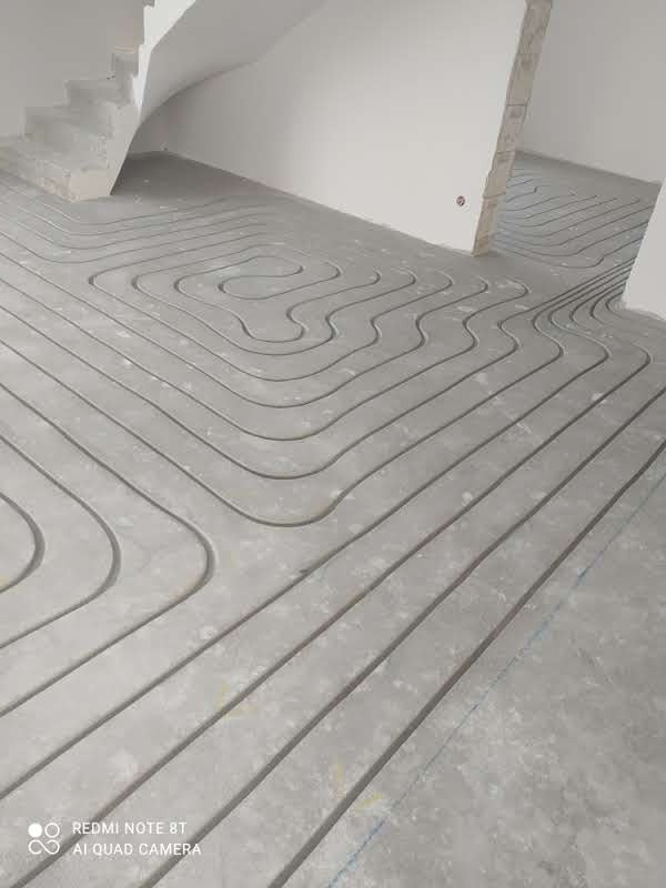 Szare rury ułożone w serpentynę na betonowej posadzce, przygotowane do zalania w systemie ogrzewania podłogowego, widoczne fragmenty schodów i surowych ścian.