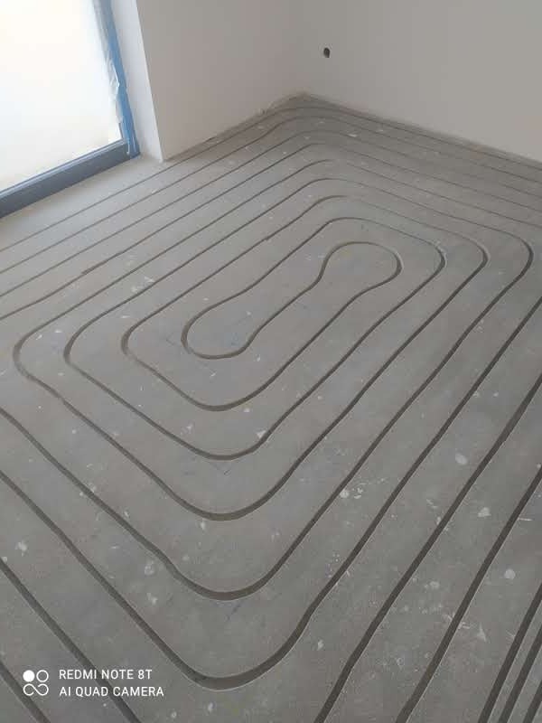 Betonowa wylewka z wyżłobionymi kanałami w kształcie pętli pod rury systemu grzewczego podłogowego, widok z góry na fragment podłogi w surowym stanie przed położeniem warstwy wykończeniowej.