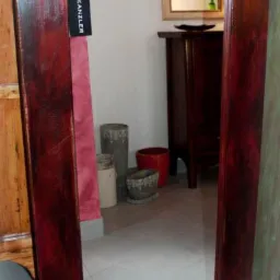 Wysokie lustro w szerokiej, ciemnoczerwonej ramie z efektem postarzenia, z widoczną metką producenta, odbijające fragment wnętrza z komodą i mniejszym lustrem.