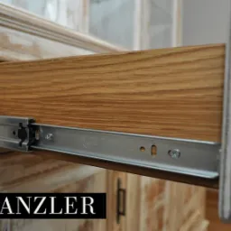 Wysunięta szuflada z jasnego drewna dębowego, widoczne metalowe prowadnice z napisem 'Versatile', front mebla w stylu rustykalnym z przetarciami farby, logo 'KANZLER' w lewym dolnym rogu.