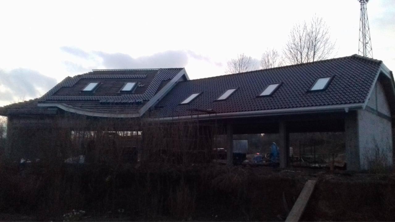 Niedokończony budynek z ciemną dachówką i oknami dachowymi, widoczny zarośnięty teren i maszt telekomunikacyjny w tle.