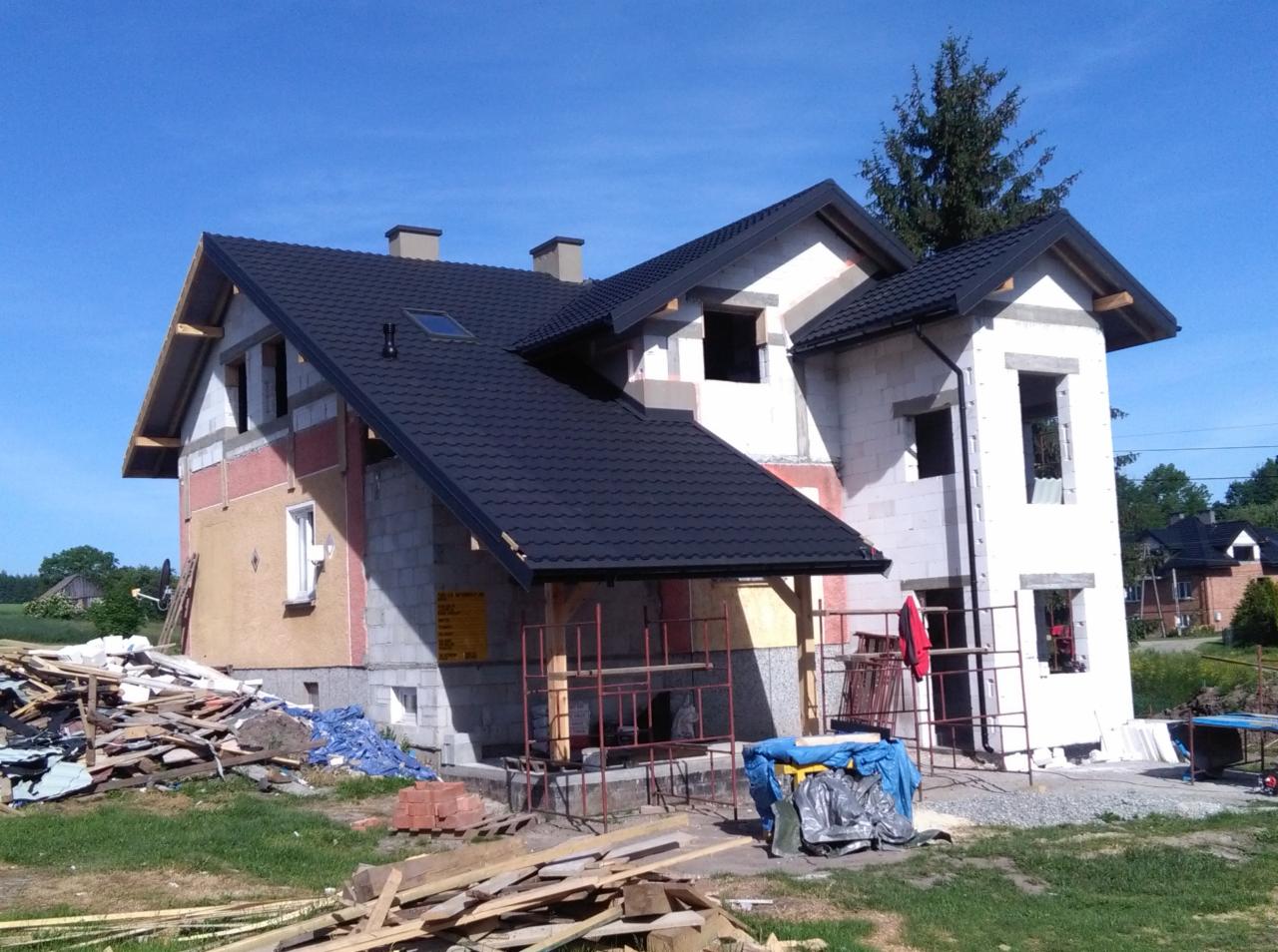 Dom w trakcie budowy z zamontowanym czarnym dachem z blachodachówki, widoczne rusztowania i materiały budowlane na trawniku.