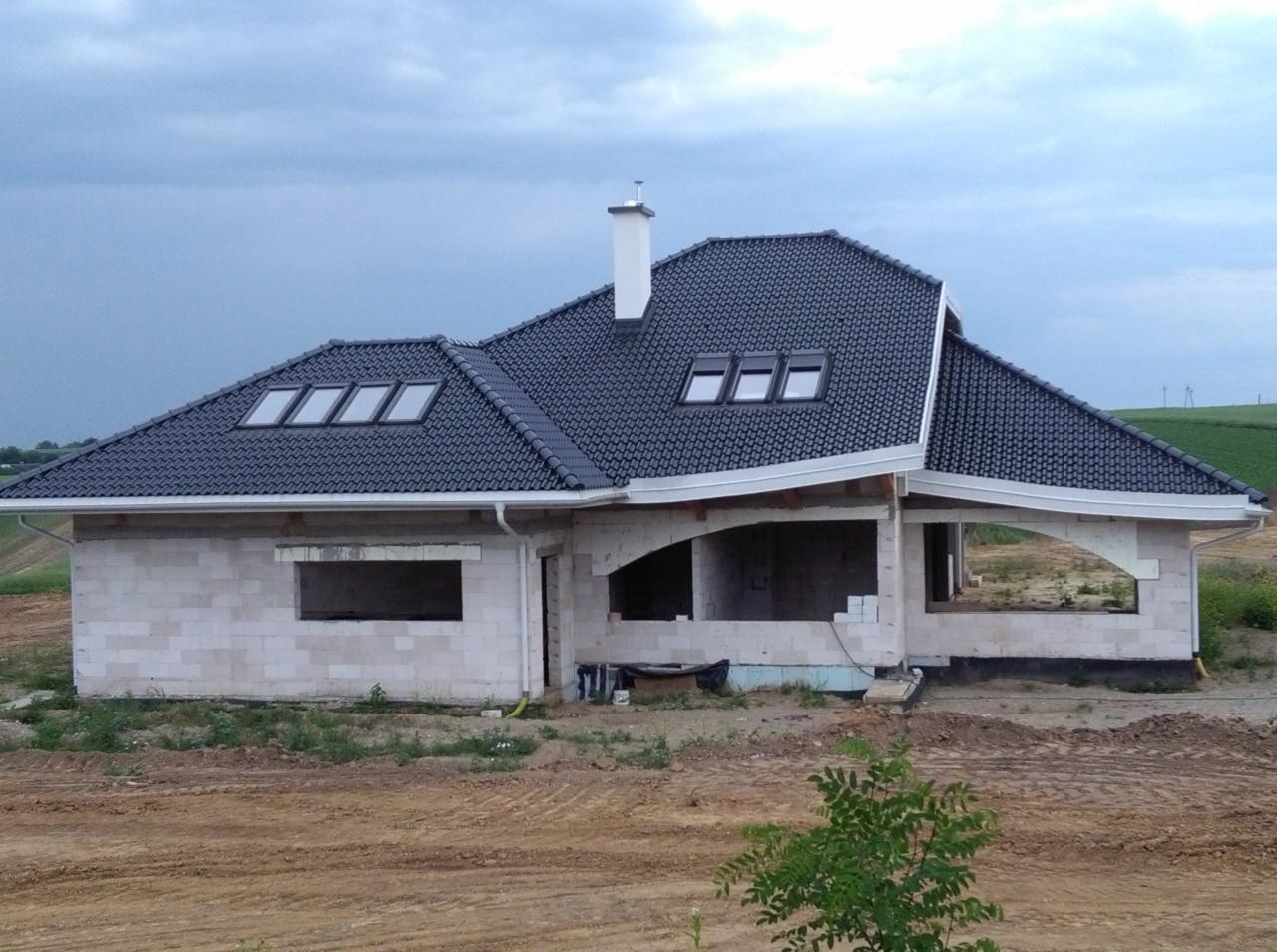 Dom w trakcie budowy z ciemną dachówką, białym kominem i rynnami. Widoczne okna dachowe i niezabudowane otwory okienne. Tło stanowi zachmurzone niebo i pole uprawne.