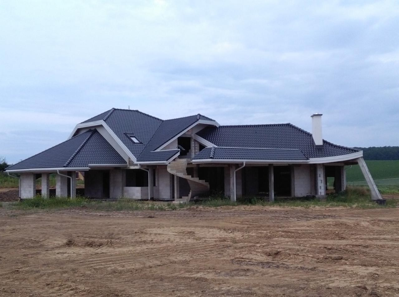 Dom jednorodzinny w trakcie budowy z ciemną dachówką, spiralnymi schodami zewnętrznymi i widocznymi elementami konstrukcyjnymi.