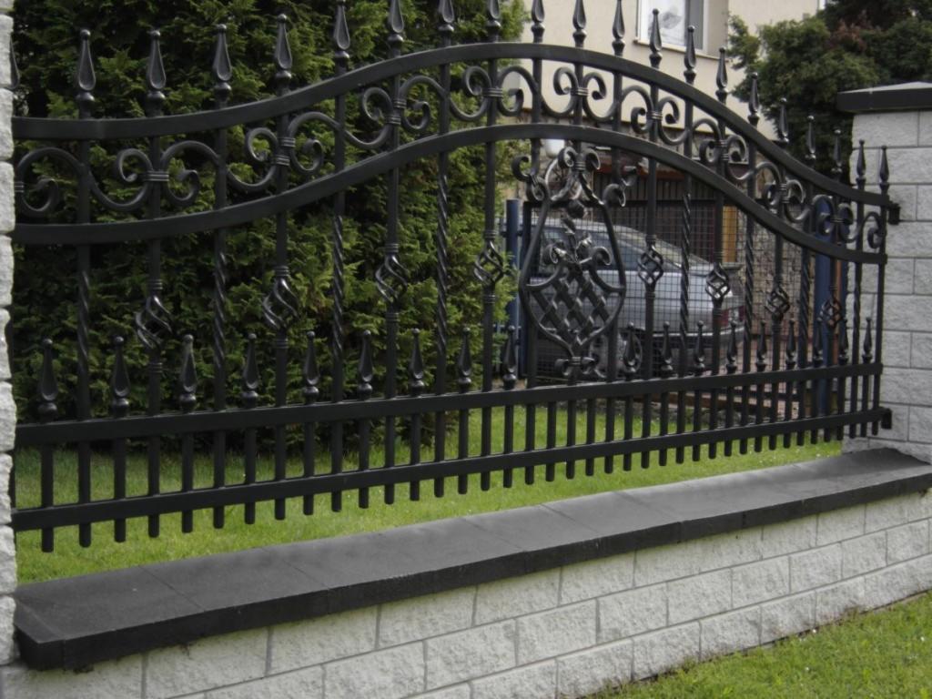 Eleganckie, czarne ogrodzenie metalowe z ozdobnymi elementami, osadzone na niskim, murowanym cokole. W tle zielony żywopłot i fragment budynku.