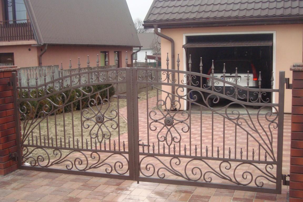 Kuta, brązowa brama wjazdowa z dekoracyjnymi elementami na tle domu z garażem, widoczna z bliska na tle kostki brukowej.