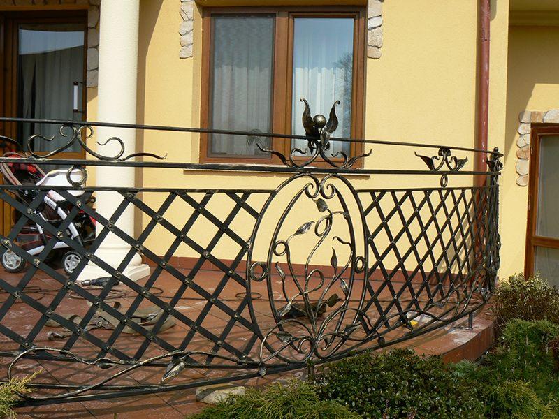 Kuty balkon z motywami roślinnymi, czarna metaloplastyka na tle żółtej elewacji domu z oknem. Widoczne detale zdobień i fragment wózka dziecięcego.