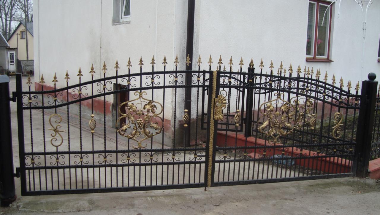Kuta, czarna brama wjazdowa z dekoracyjnymi, złotymi elementami na tle białego budynku. Brama dwuskrzydłowa, z ornamentami roślinnymi i geometrycznymi.