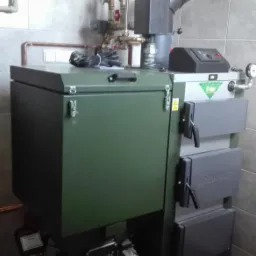 Nowoczesny, zielono-szary piec na paliwo stałe marki DREW-MET z widocznymi elementami instalacji, w tym pompami obiegowymi, zamontowany w kotłowni wyłożonej płytkami ceramicznymi.