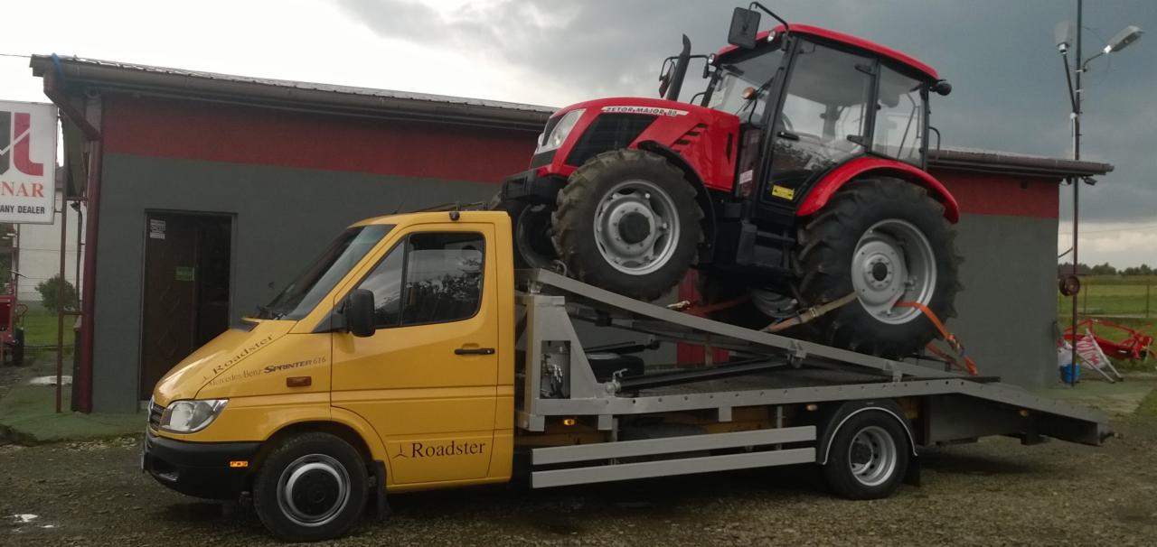 Żółta laweta Mercedes-Benz Sprinter 616 przewozi czerwony traktor Zetor Major 80 na tle budynku. Transport specjalistyczny.