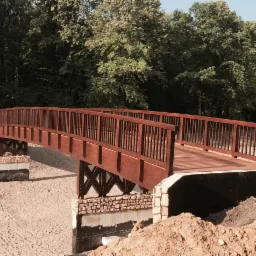 Drewniany most łukowy z brązowymi barierkami, wsparty na betonowych i kamiennych podporach, w otoczeniu piasku i drzew.