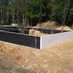 Wykop pod fundamenty domu z hydroizolacją pionową w kolorze czarnym i ociepleniem ze styropianu, widoczne zbrojenia pionowe i pracownik w tle.