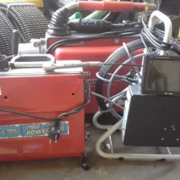 Sprzęt do inspekcji kanalizacji: kamera inspekcyjna z monitorem i urządzenie czyszczące z elastycznym wężem na szpuli. Widoczne detale: etykiety na urządzeniach, przewody i wtyczka zasilająca.