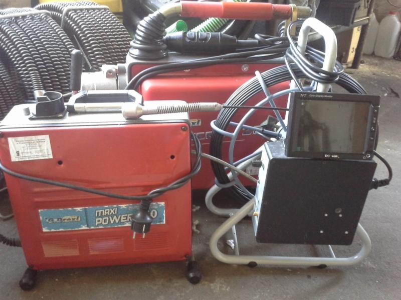 Sprzęt do inspekcji kanalizacji: kamera inspekcyjna z monitorem i urządzenie czyszczące z elastycznym wężem na szpuli. Widoczne detale: etykiety na urządzeniach, przewody i wtyczka zasilająca.