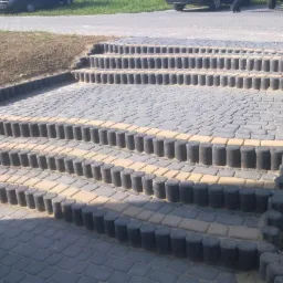 Kamienne schody z kostki brukowej w odcieniach szarości i beżu, obrzeża z ciemnych, betonowych słupków. Widok z góry, oświetlenie dzienne.