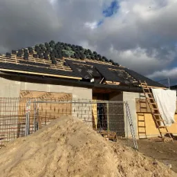 Budowa domu w trakcie: czarne dachówki układane na łatach na nowym budynku, widoczna sterta piasku i drabina oparta o ścianę.