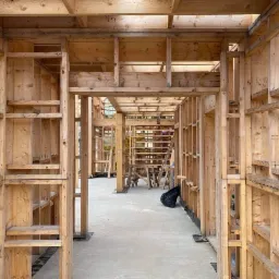 Surowa drewniana konstrukcja domu w trakcie budowy, widoczne liczne drewniane belki i słupy tworzące szkielet budynku, betonowa podłoga, w tle drabina i inne elementy konstrukcji.