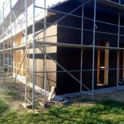 Dom w trakcie ocieplania elewacji zewnętrznej, widoczne rusztowania i czarne płyty izolacyjne na ścianach, okna bez szyb.