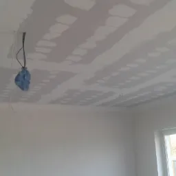 Sufit z płyt kartonowo-gipsowych po szpachlowaniu, widoczne łączenia. Kabel zwisający z sufitu zabezpieczony niebieską folią. W tle okno z widokiem na zieleń.