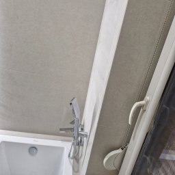 Hubert Szulc wykończenia Wnetrz - Jasna łazienka po remoncie: biała wanna, chromowana bateria, beżowe płytki i okno z roletą. Minimalistyczny design, czyste linie, nowoczesne wykończenie.