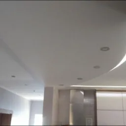 Biały sufit podwieszany z wbudowanymi punktowymi lampami LED i dekoracyjnym zakrzywionym wgłębieniem oświetlonym od góry, widok z dołu na fragment salonu i kuchni.