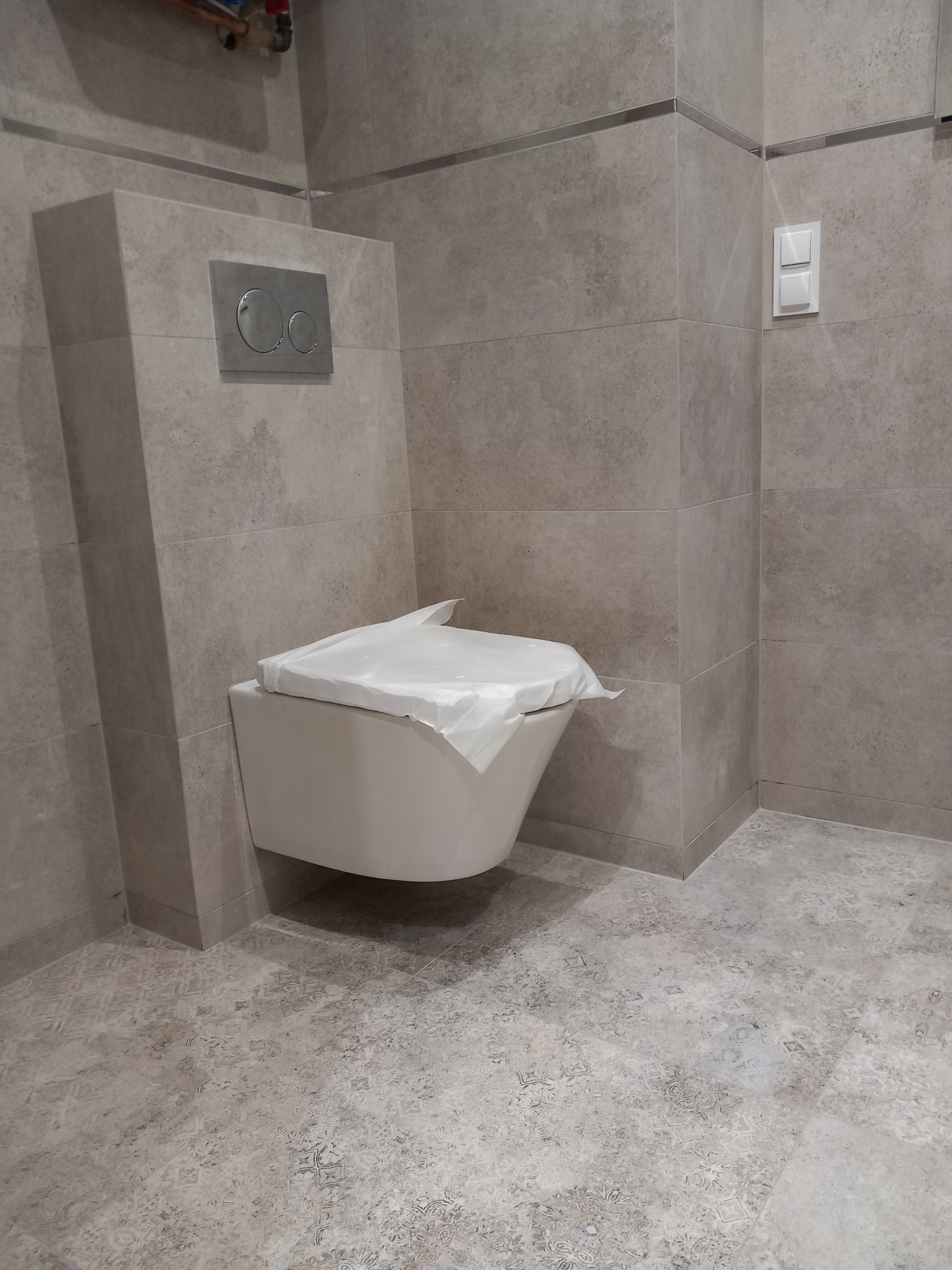 Nowoczesna, podwieszana toaleta w łazience wykończonej w szarej kolorystyce, z częściowo zakrytym siedziskiem folią ochronną, osadzona na tle ściany wyłożonej płytkami imitującymi kamień...