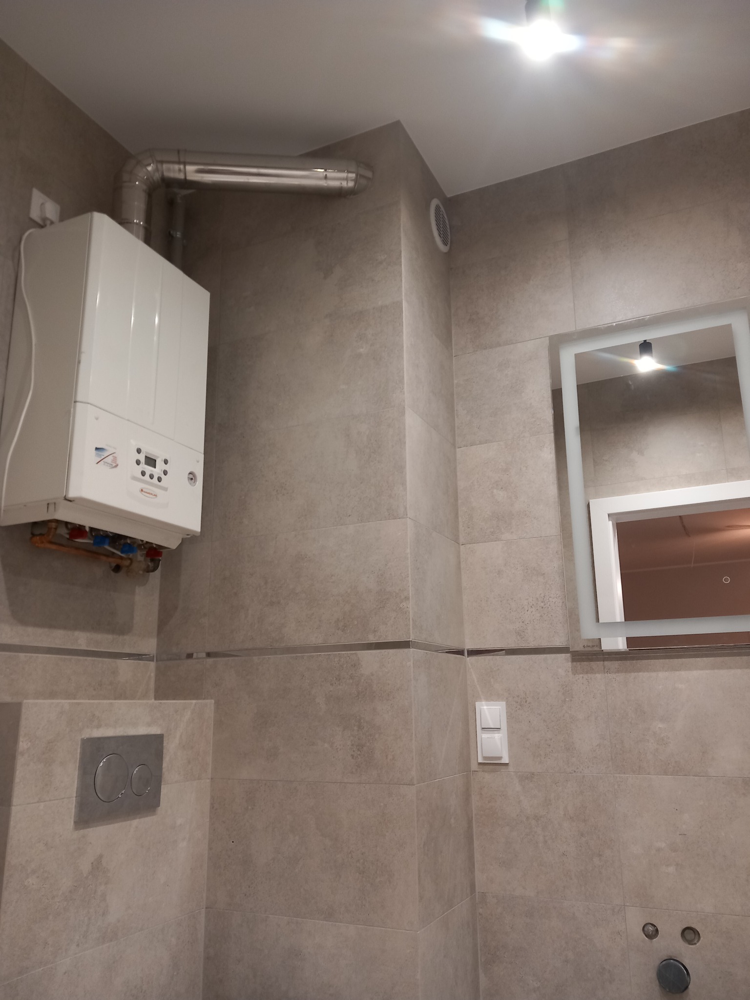 Kompaktowa łazienka z beżowymi płytkami, wiszącym kotłem gazowym z odprowadzeniem spalin, podświetlanym lustrem i przyciskiem spłuczki WC.