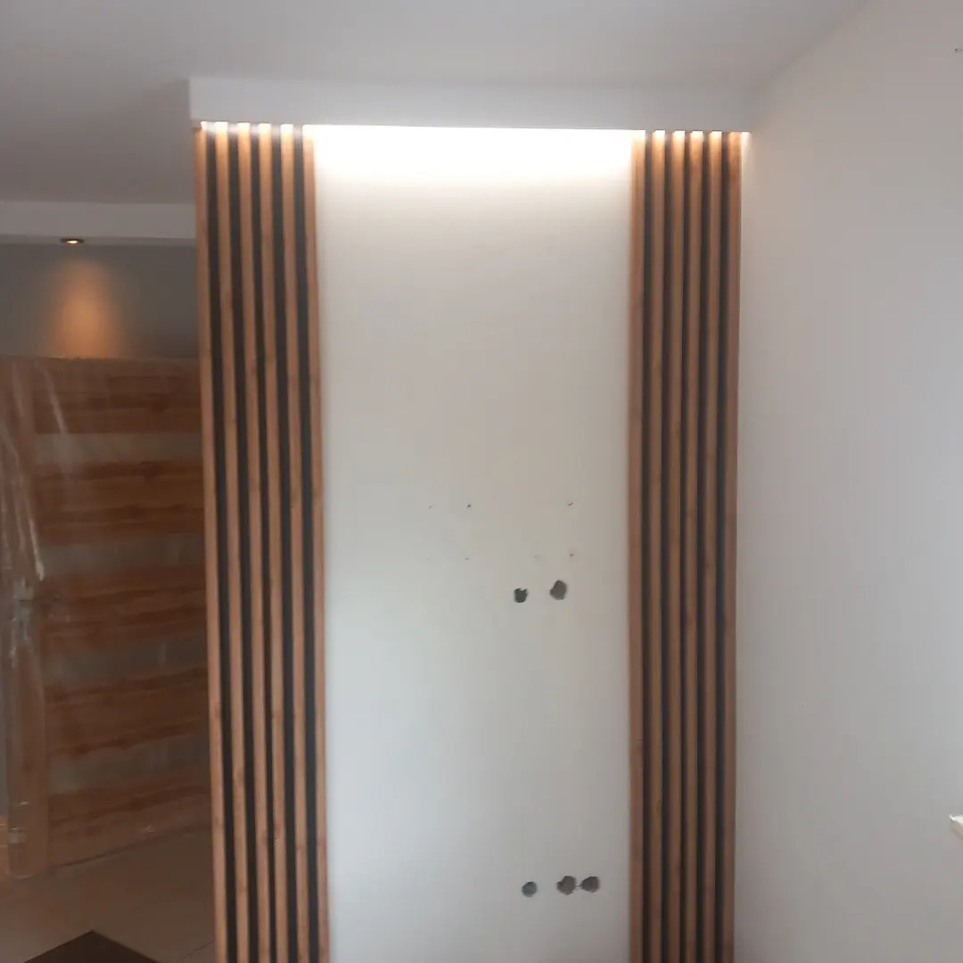 Ściana z drewnianymi lamelami po bokach i podświetleniem LED, widoczne otwory po gniazdkach i ślady po wierceniu, w tle drzwi zabezpieczone folią.