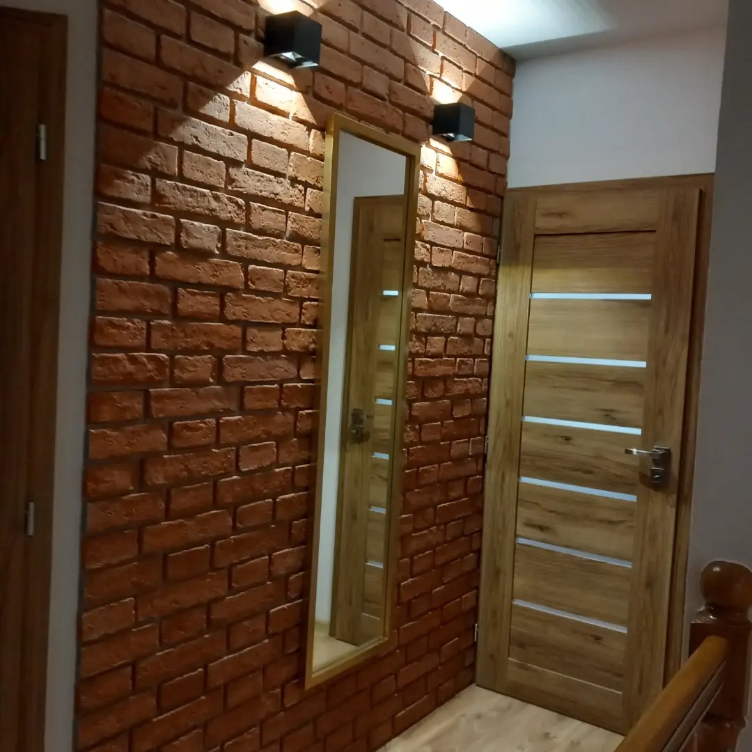 Ściana z cegły klinkierowej w korytarzu z dwoma czarnymi kinkietami, lustrem w złotej ramie i drewnianymi drzwiami z poziomymi przeszkleniami.