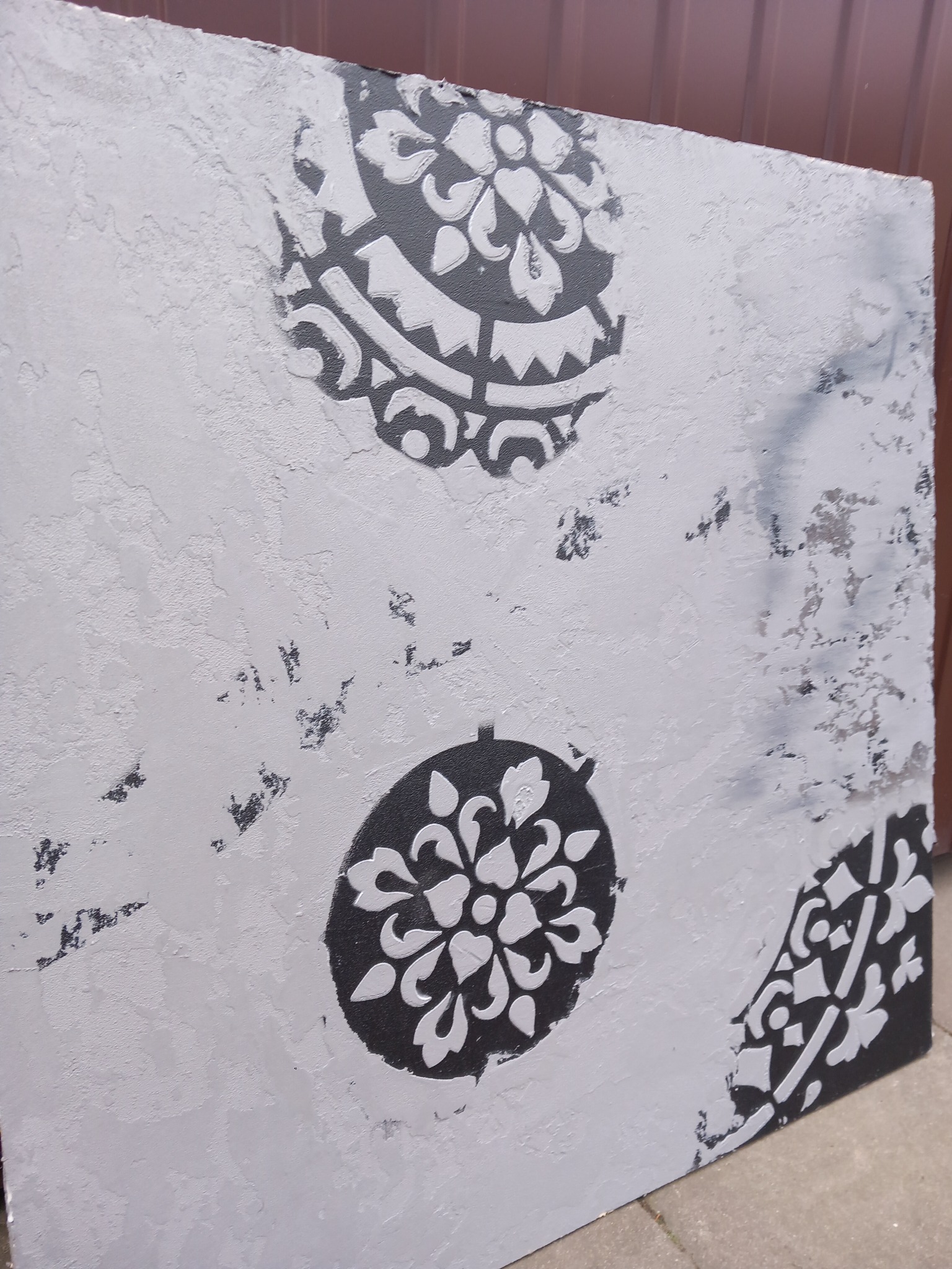 Szara ściana z dekoracyjnym, czarnym wzorem w kształcie rozet i ornamentów, wykonana techniką szablonową.