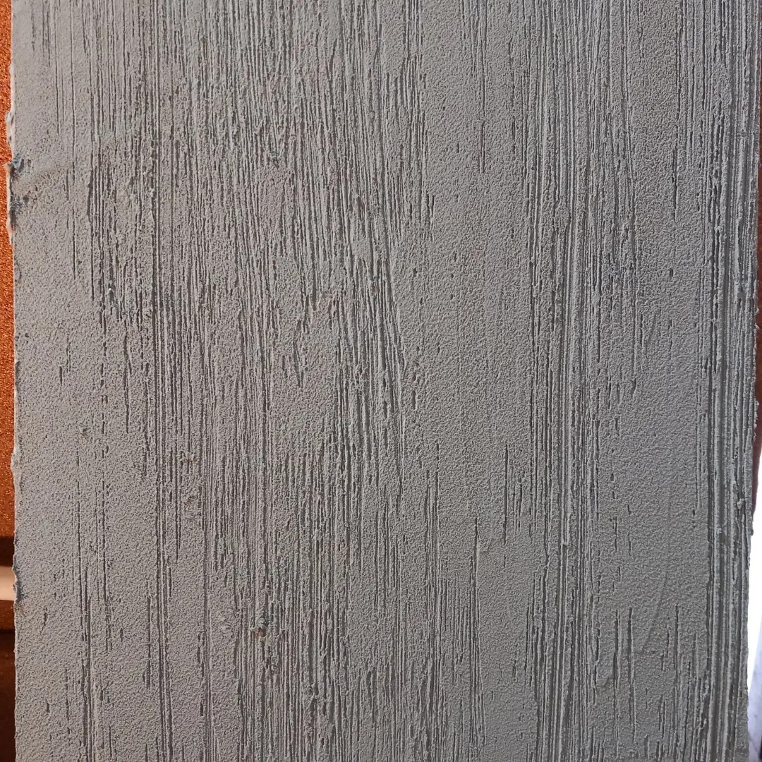 Szara ściana z wyraźną, pionową fakturą, przypominającą drapanie, z widocznym fragmentem pomarańczowego narożnika po lewej stronie.