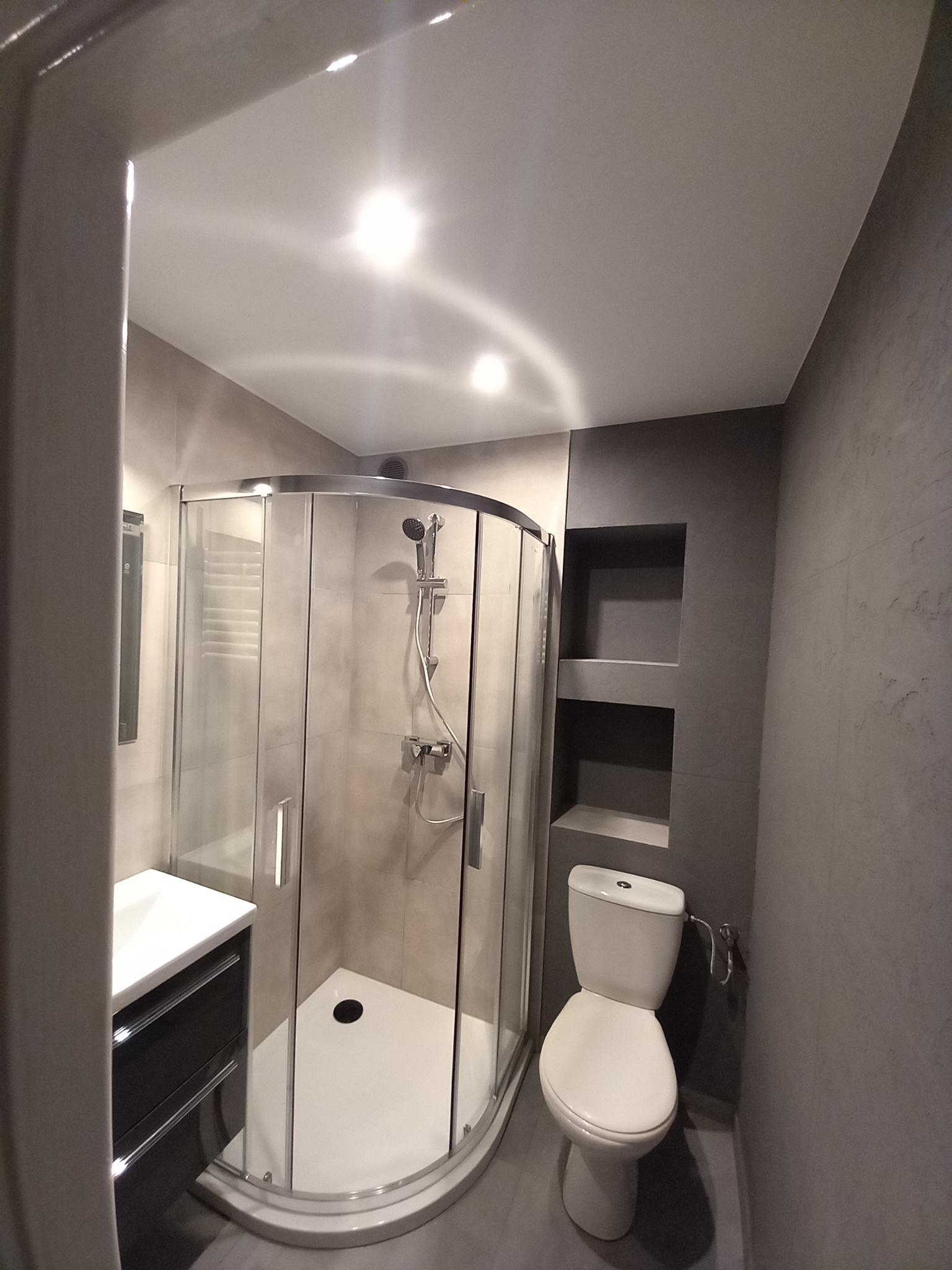 Nowoczesna łazienka z kabiną prysznicową typu walk-in, umywalką z szafką w kolorze grafitowym, toaletą i wnękami w ścianie, wykończona w odcieniach szarości.