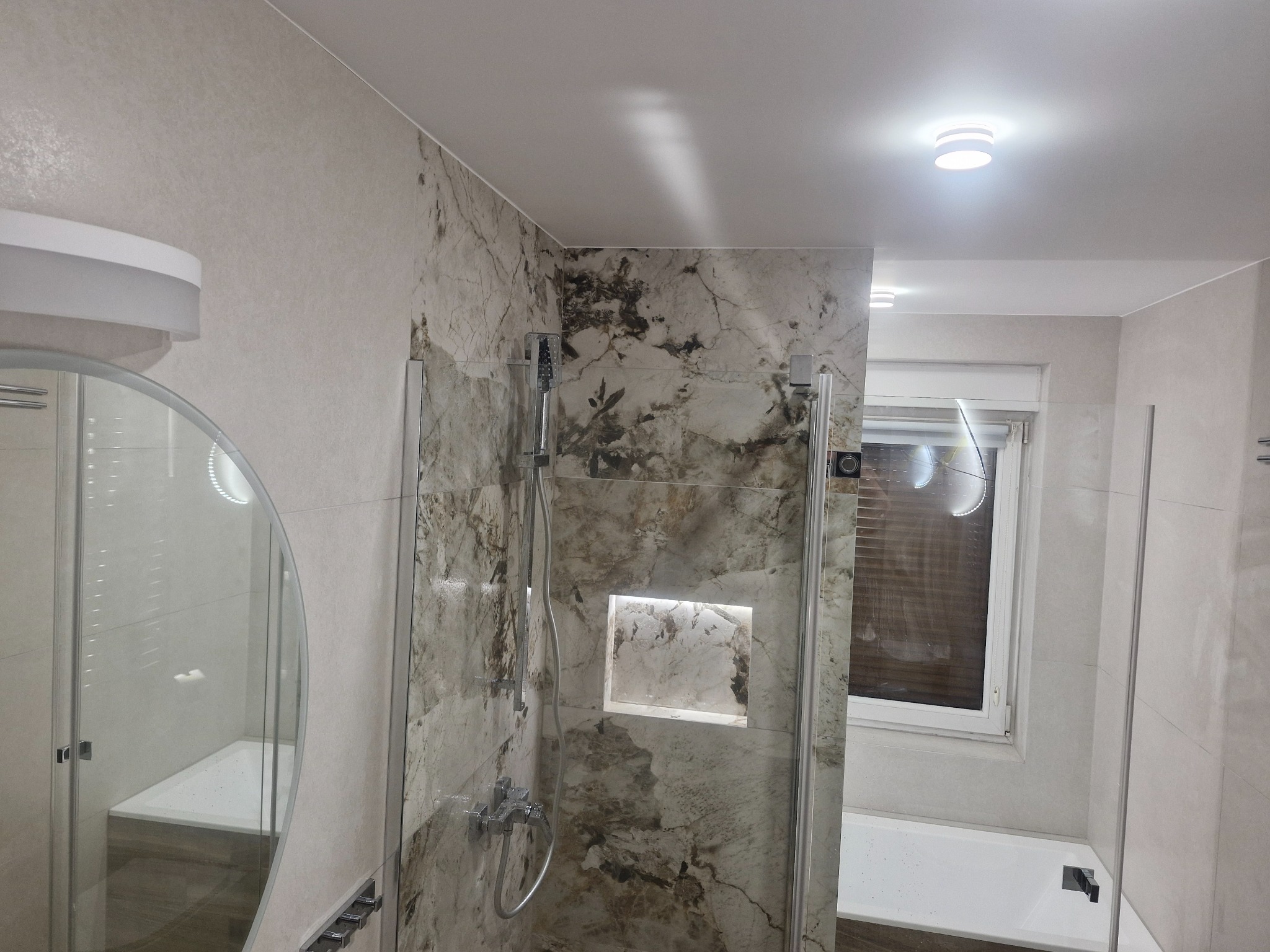 Wykończona łazienka z kabiną prysznicową typu walk-in, wyłożona płytkami imitującymi marmur. Widoczne oświetlenie sufitowe i okrągłe lustro.