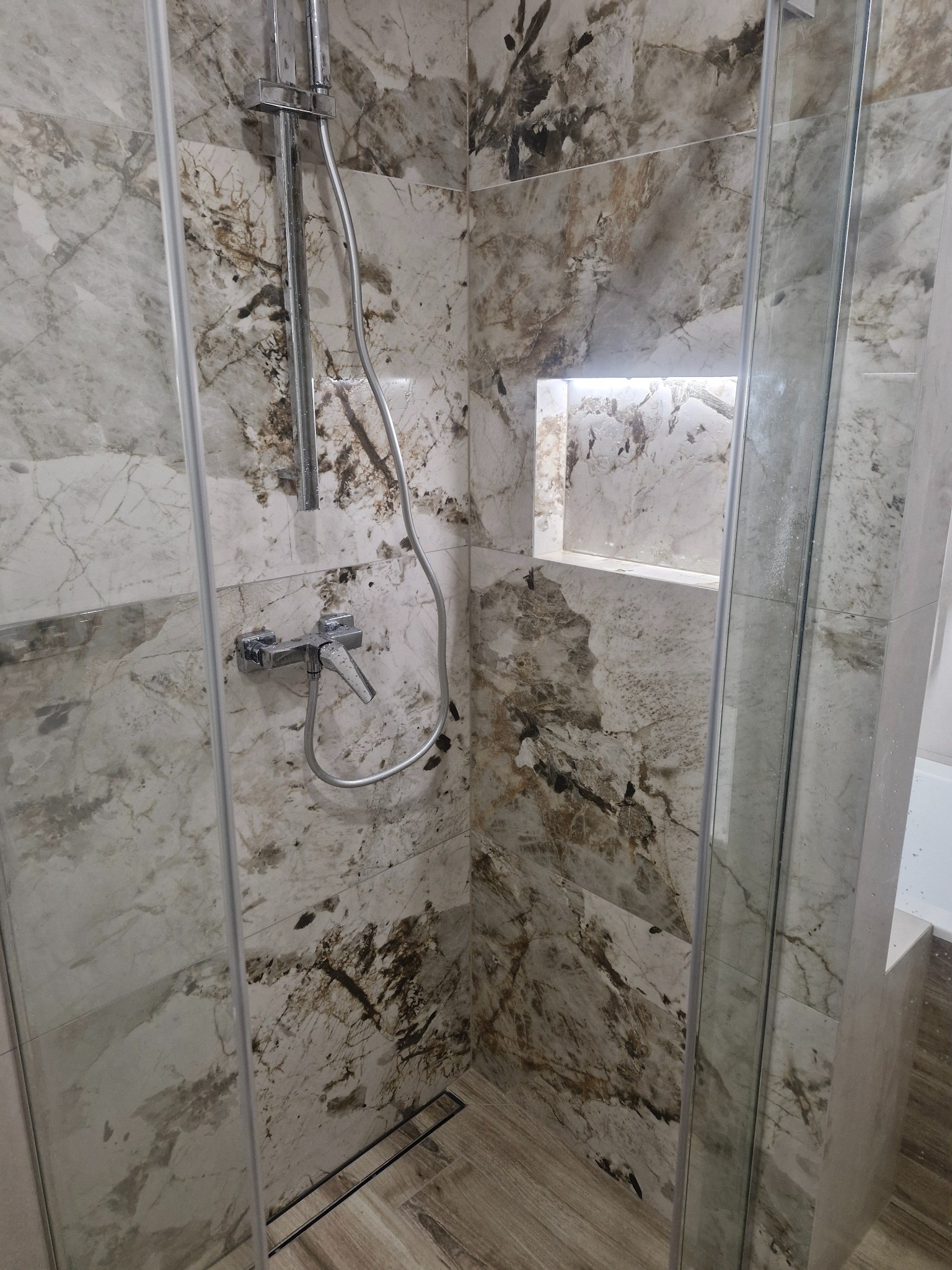 Kabina prysznicowa z marmurowymi płytkami, chromowaną armaturą i odpływem liniowym. W ścianie wnęka z podświetleniem LED. Widoczne detale wykończenia.