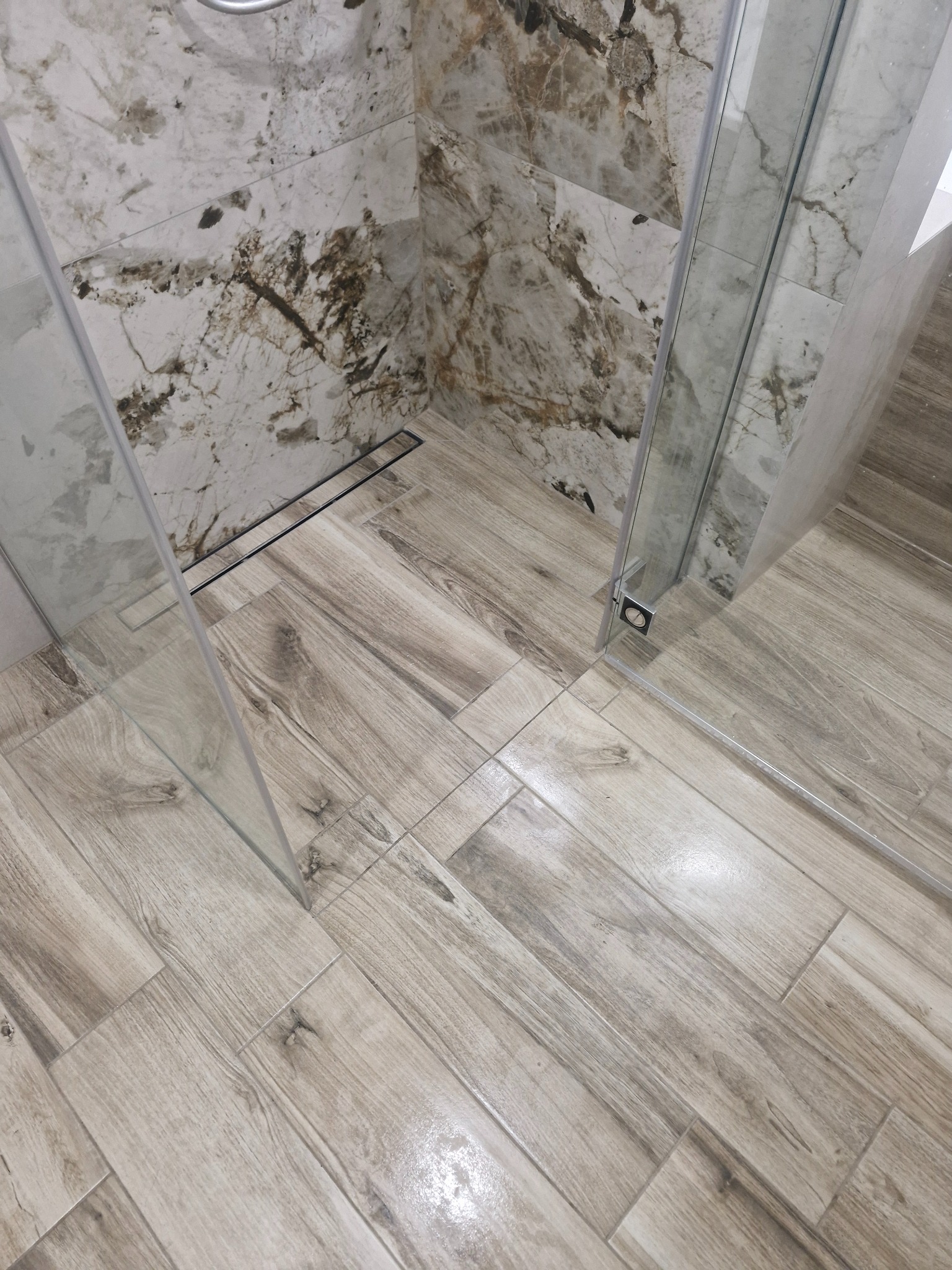 Wykończona kabina prysznicowa z marmurowymi płytkami ściennymi i drewnopodobną podłogą. Widoczny odpływ liniowy i szklane drzwi kabiny.