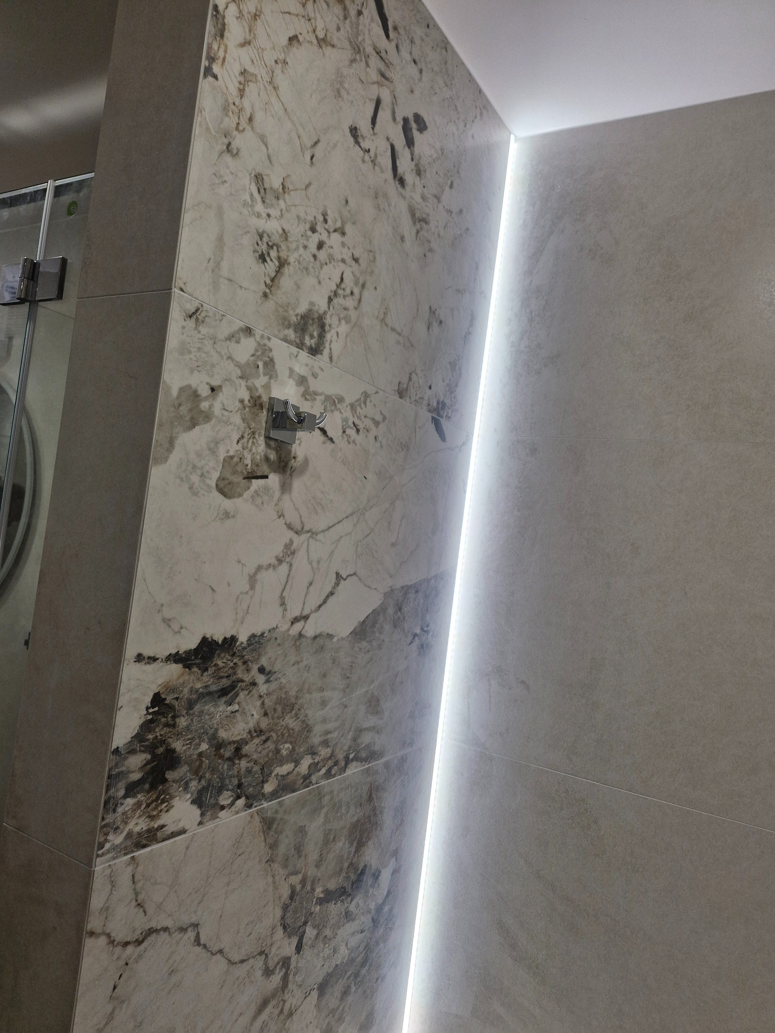 Wykończona łazienka: ściana z płytek imitujących marmur z oświetleniem LED, widoczny fragment kabiny prysznicowej. Nowoczesny design i precyzyjne wykonanie.