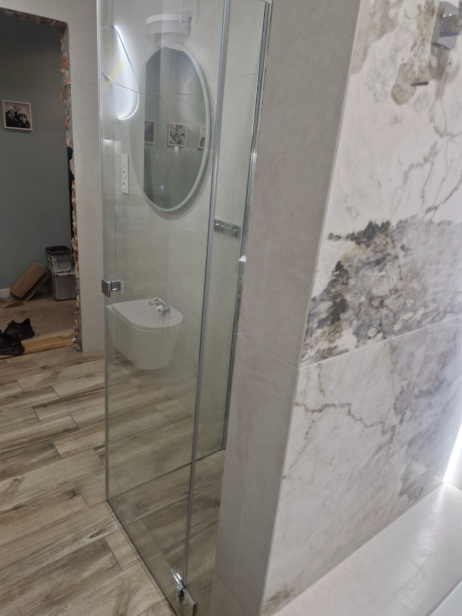 Wykończona łazienka z kabiną prysznicową, bidetem i podświetlanym lustrem. Widoczne płytki imitujące drewno na podłodze i marmur na ścianach. Częściowo widoczny remont.