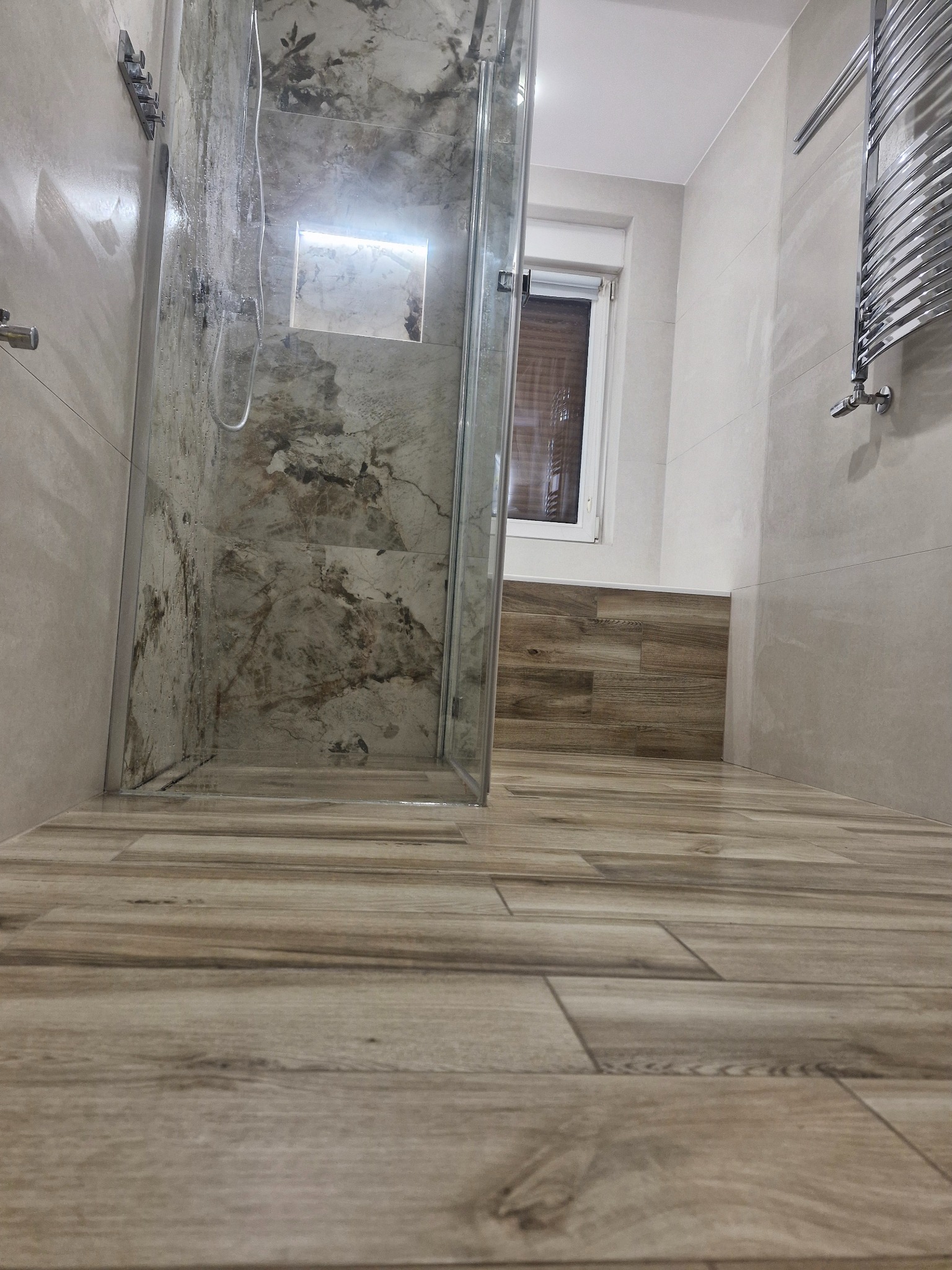 Nowoczesna łazienka z kabiną prysznicową typu walk-in, wykończona płytkami imitującymi drewno i marmur. Widoczny grzejnik drabinkowy i okno.