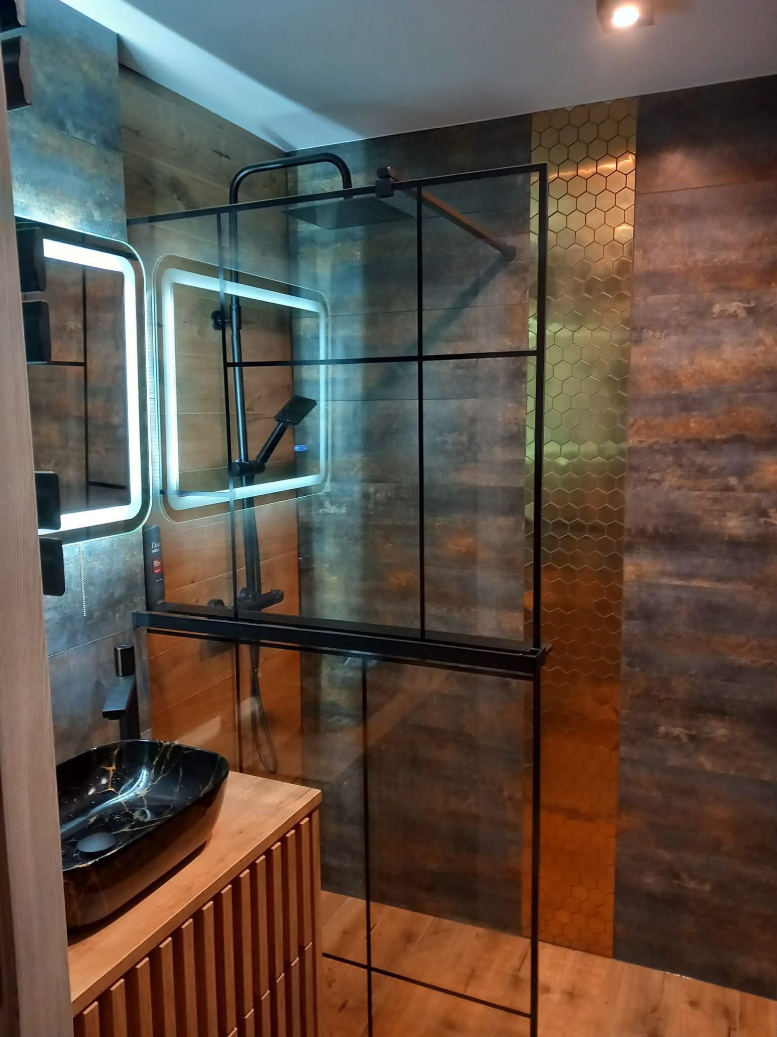 Nowoczesna łazienka z kabiną prysznicową typu walk-in, umywalką nablatową w kolorze czarnym i podświetlanym lustrem. Na ścianie dekoracyjne płytki w odcieniach brązu i złota.