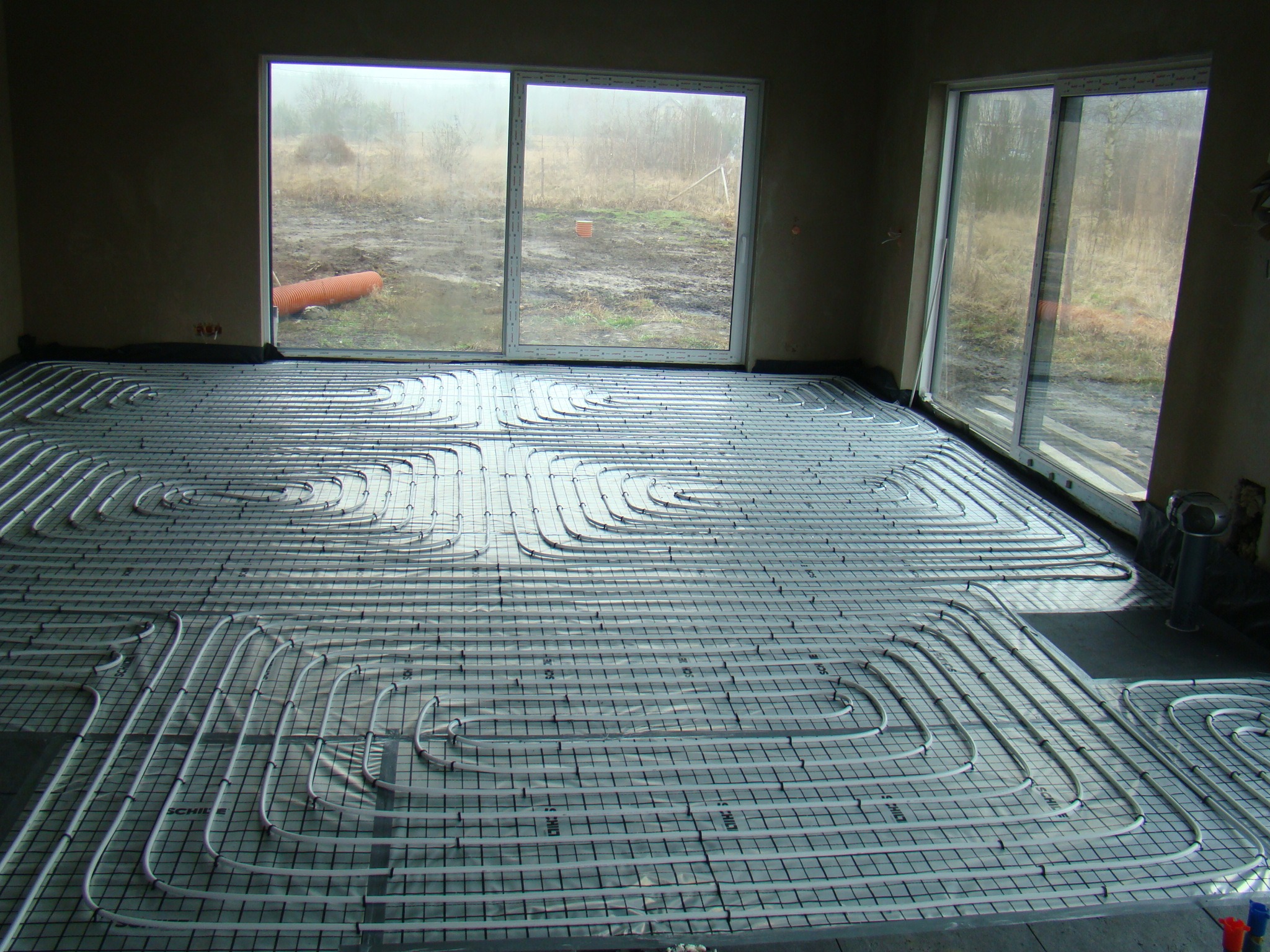 Rozłożone rury ogrzewania podłogowego na folii z rastrem w surowym pomieszczeniu z dużymi oknami wychodzącymi na zewnątrz w pochmurny dzień.