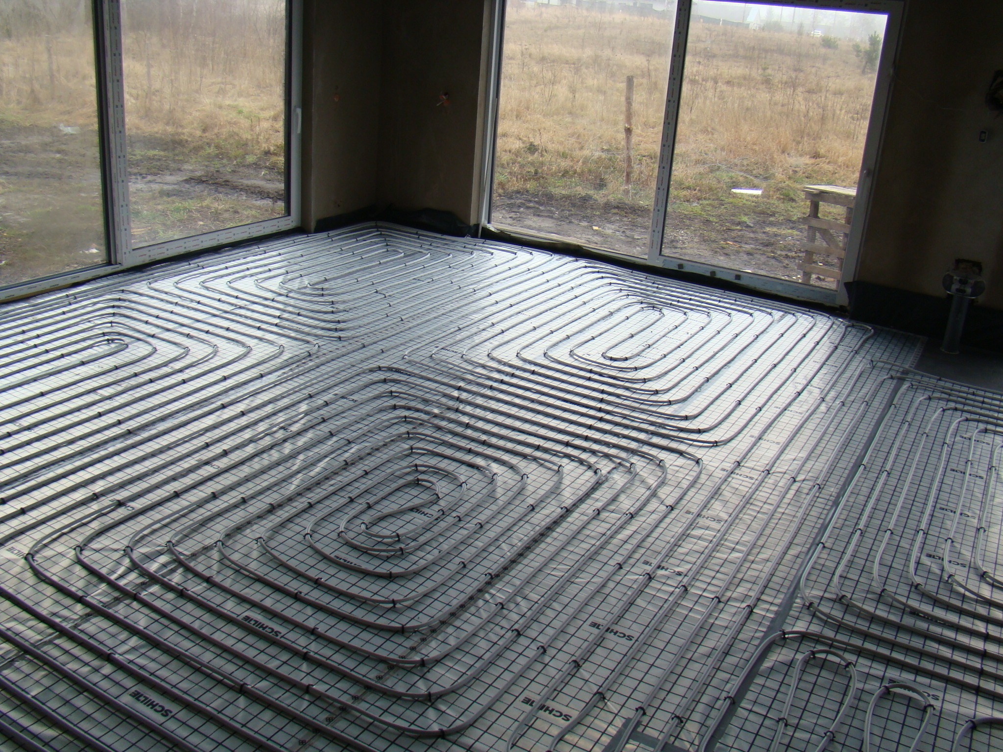 Rury ogrzewania podłogowego ułożone na siatce w surowym wnętrzu z dużymi oknami wychodzącymi na zewnątrz.