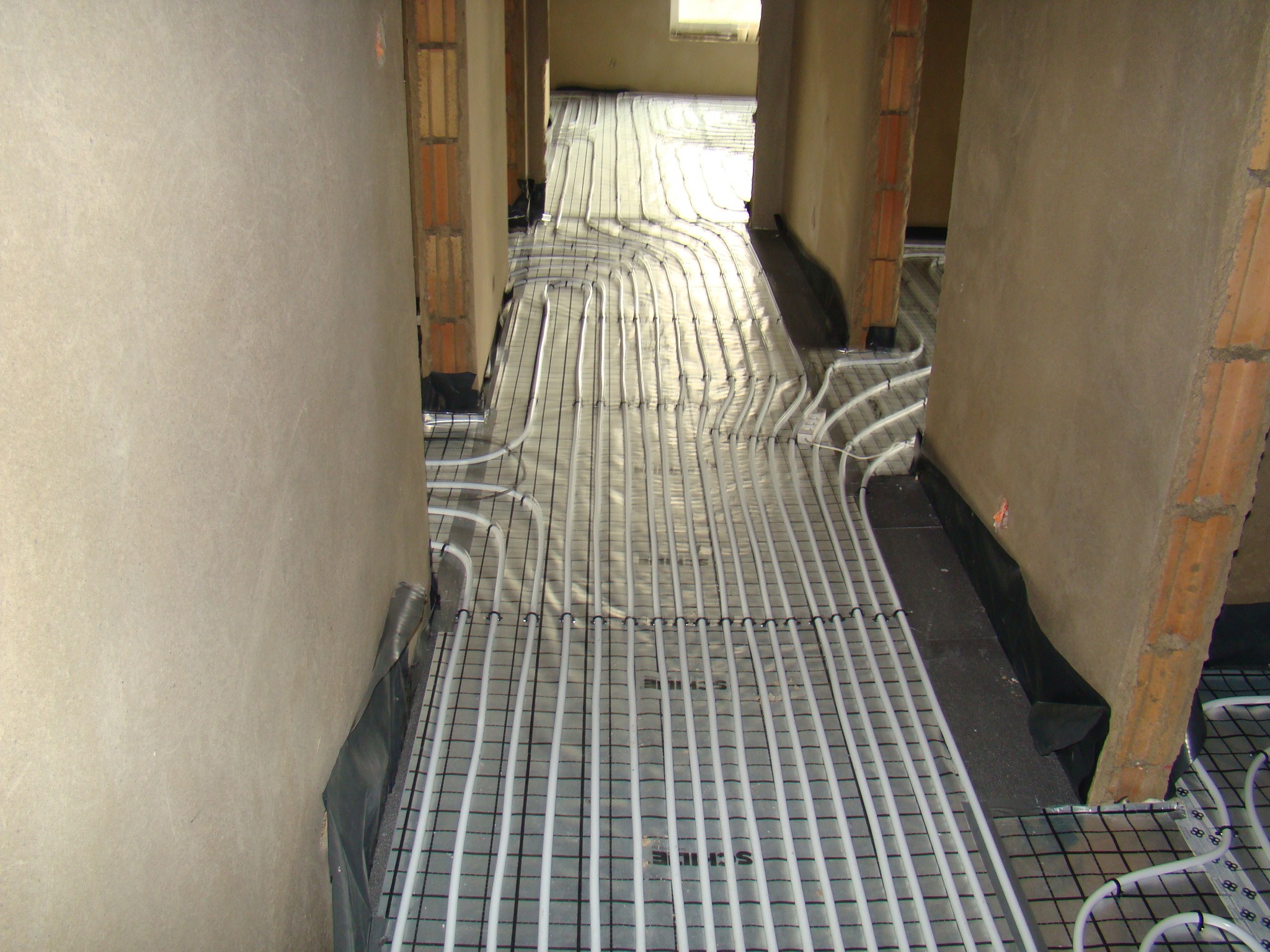 Rozłożone rury ogrzewania podłogowego na folii izolacyjnej z siatką, widok wzdłuż korytarza w surowym stanie.