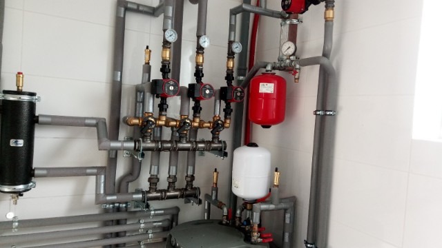 Złożona instalacja hydrauliczna z czterema pompami obiegowymi, zbiornikiem wyrównawczym i izolowanymi rurami na tle białych płytek.