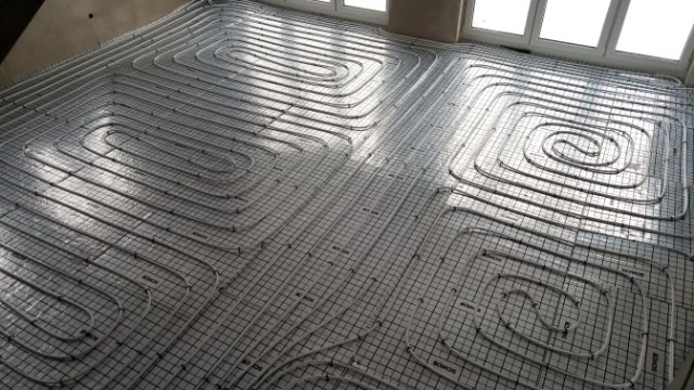 Białe rury ogrzewania podłogowego ułożone spiralnie na folii z nadrukowaną siatką, widoczne okno w tle.