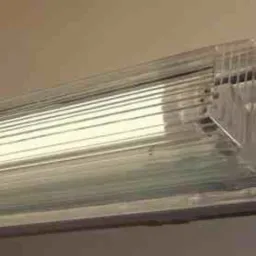 Zamknięta, podłużna lampa LED z przezroczystą obudową zamontowana na beżowej ścianie. Widoczna struktura lampy i odbijające się światło.