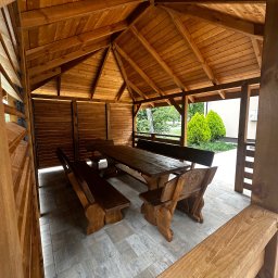 Tartak Zągoty tarasy , patio altany - Drewniana altana z masywnym stołem i ławami, widok z zewnątrz. Konstrukcja z jasnego drewna, zadaszenie skośne, częściowo zabudowane ściany, podłoga wyłożona płytkami.